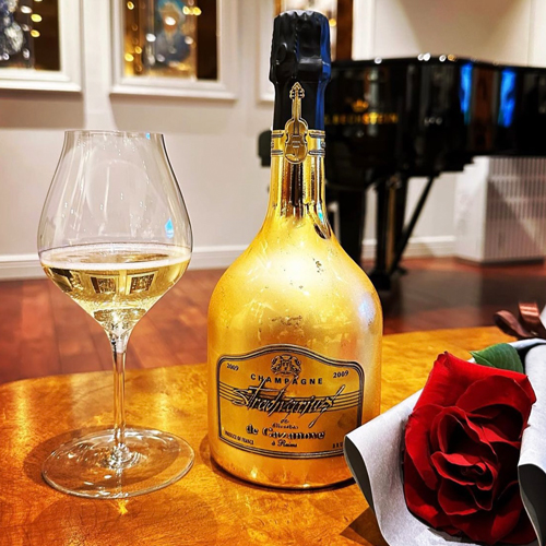une bouteille de Stradivarius Gold 2009 posée sur une table avec une coupe et une rose devant un piano / A bottle of Stradivarius Gold 2009 placed on a table with a cup and a rose in front of a piano.